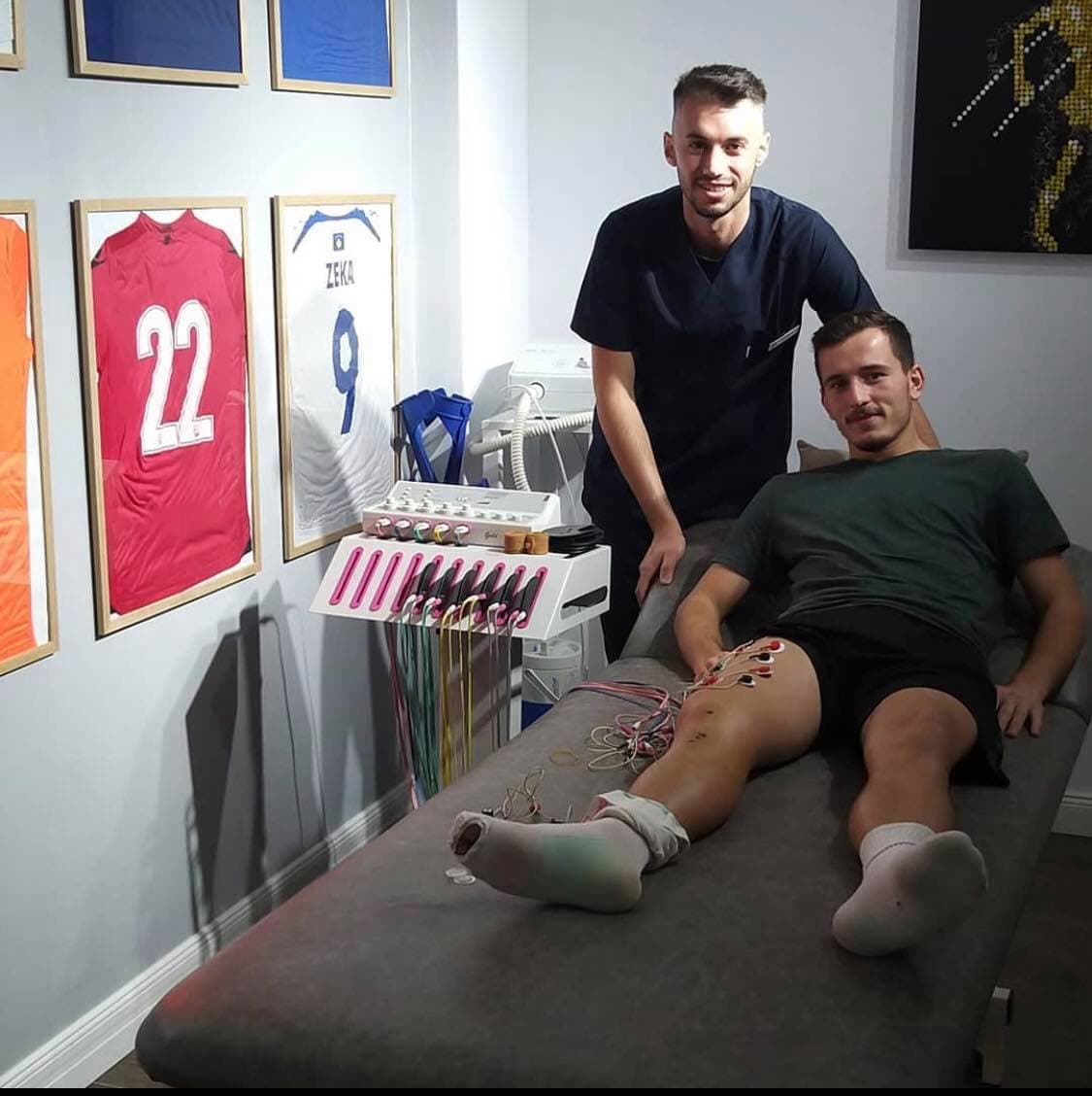 Arbanis Maxhuni pranë një futbollisti gjatë elektroterapisë për gjurin, me fanella futbolli në mur